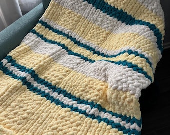 May include: A chunky knit blanket in shades of yellow, white, and teal. The blanket features horizontal stripes of color, creating a cozy and textured appearance. The blanket is draped over a teal armchair.