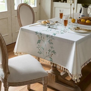 May include: A white tablecloth with a green floral pattern and tassel edging. The table is set with plates, glasses, and food. The table is made of wood and has two chairs. The tablecloth is rectangular.