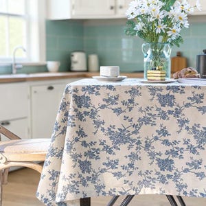 May include: A round tablecloth with a cream background and a blue floral pattern. The tablecloth is on a round table with a vase of white flowers, a cup and saucer, and a small cake. The table is in a kitchen setting.