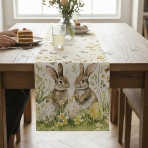 May include: A rectangular table runner featuring a watercolor design of two brown rabbits, yellow flowers, and Easter eggs. The runner is placed on a wooden table, creating a spring or Easter theme.