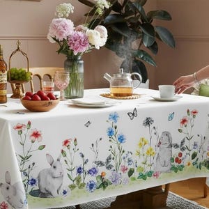 Puede incluir: Un mantel blanco con un diseño floral y de conejos. El mantel presenta flores coloridas, mariposas y conejos. Una tetera, una taza de té y un jarrón de flores están sobre la mesa. También hay una botella de licor y un cuenco de fresas.