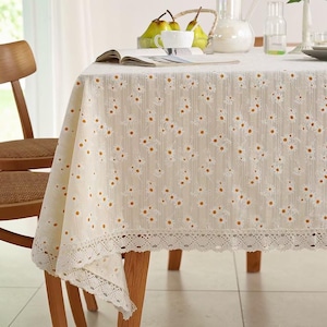 Könnte beinhalten: Cremefarbene Tischdecke mit Gänseblümchenmuster und Spitzenbesatz. Die Tischdecke liegt auf einem Holztisch. Ein Buch, Birnen und eine Tasse befinden sich auf dem Tisch. Ein Holzstuhl ist im Hintergrund zu sehen.