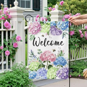 Könnte beinhalten: Eine weiße Gartenflagge mit dem Wort "Welcome" in schwarzer Schrift, umgeben von bunten Hortensienblüten in Rosa, Blau, Lila und Grün. Zwei kleine Bienen sind ebenfalls im Design enthalten. Die Flagge wird im Freien gezeigt.