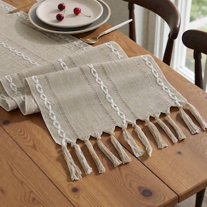 Könnte beinhalten: Beiger Tischläufer aus Leinen mit weißen Stickereidetails und Fransenquasten. Der Läufer liegt auf einem Holztisch, im Hintergrund sind ein Teller und Besteck zu sehen. Ein dekoratives Element für das Esszimmer.