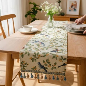 May include: A wooden dining table with a decorative bird and floral patterned table runner. The runner has a cream background with blue birds, green leaves, and yellow flowers. A stack of plates is on the table.