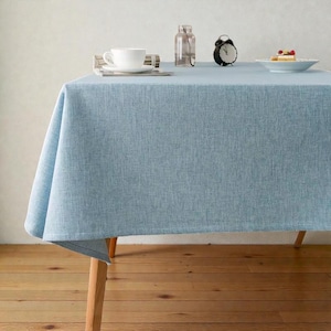 May include: A pale blue linen tablecloth on a wooden table. A white teacup and saucer, a small black alarm clock, and a plate with a dessert are on the table. A clear glass bottle sits nearby.