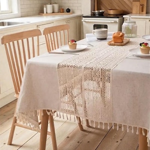 Peut inclure: Une nappe en lin beige avec un chemin de table en dentelle crème. Le chemin de table présente un motif ajouré détaillé et est frangé aux extrémités. Deux chaises en bois sont visibles, ainsi qu'une table avec des cupcakes et un petit pain.