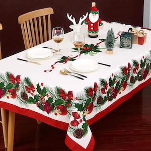 May include: A white Christmas tablecloth with a red border, decorated with green pine branches, red bows, and pinecones. The table is set with white plates, gold and black cutlery, and wine glasses. A Santa bottle cover and decorative reindeer are also on the table.