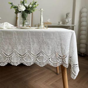 May include: A white rectangular tablecloth featuring intricate floral embroidery and a scalloped lace border. The fabric has a textured finish and hangs over the edges of a wooden table, showcasing detailed needlework patterns along the hem.