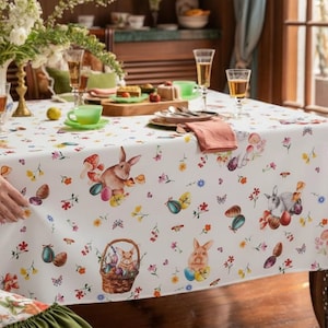 May include: White Easter tablecloth with illustrations of bunnies, eggs, flowers, and butterflies. The table is set with champagne flutes, a vase of white flowers, and a wooden serving board. A hand is seen adjusting the tablecloth.