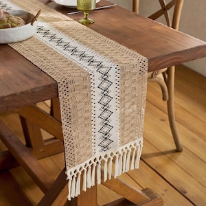 May include: A woven beige table runner with a geometric design in black, white, and gray. The runner has a fringed edge and is displayed on a wooden table. The table is set with a bowl, plate, and glass.