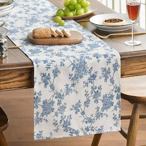 Op de afbeelding: Witte tafelloper met een blauw bloemmotief, op een houten tafel. De loper heeft een herhalend patroon van bloemen en ranken. De tafel is gedekt met borden, een wijnglas en een houten serveerplank.