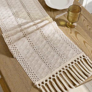 Può includere: Runner da tavolo color crema, finemente tessuto con un motivo geometrico e dettagli con frange. Il runner è posto su un tavolo di legno chiaro, accanto a un piatto bianco e un bicchiere strutturato.