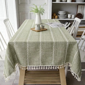 May include: A sage green rectangular tablecloth with white vertical stripes and decorative stitching. The tablecloth has a fringe of white tassels along the edges. A wooden table is set with a white vase and small ceramic items.