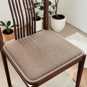 May include: A brown wooden chair with a woven beige seat cushion. The chair has a curved back with vertical slats. The cushion has a textured pattern and a light brown border. The chair is in a well-lit room with plants in the background.
