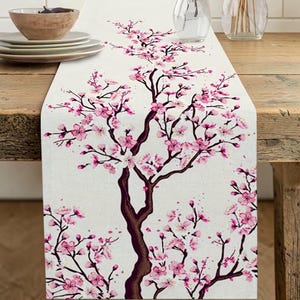 May include: A white table runner featuring a watercolor illustration of a cherry blossom tree with pink flowers and brown branches. The runner is placed on a wooden table, with plates and a bowl visible in the background.