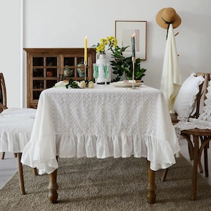 May include: A white, eyelet lace tablecloth with a ruffled edge covers a wooden table. The table is set with candles, flowers, and glassware. A matching tablecloth covers a nearby chair. A hat and towel hang on a wooden rack.