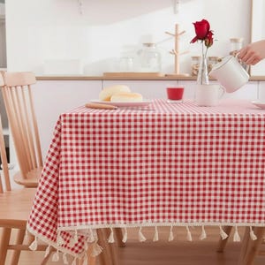 Könnte beinhalten: Eine rot-weiß karierte Tischdecke mit Quastenbesatz bedeckt einen Tisch. Zwei Holzstühle befinden sich im Vordergrund. Eine rote Rose in einer Vase und ein weißer Krug stehen auf dem Tisch. Essen und Getränke sind ebenfalls vorhanden.
