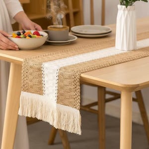 Può includere: Un tavolo da pranzo in legno chiaro con un runner da tavolo intrecciato beige e bianco. Il runner ha un bordo con frange. Un vaso bianco con verde, una ciotola di frutta e una tazza di caffè sono sul tavolo.