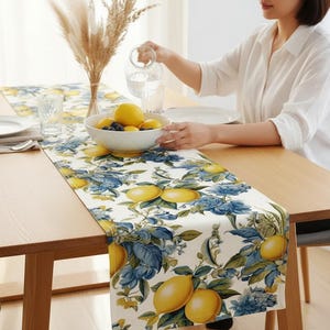 May include: A wooden dining table set with a floral table runner featuring lemons and blue flowers. A bowl of lemons and blueberries sits on the table. A person is pouring water into a glass.