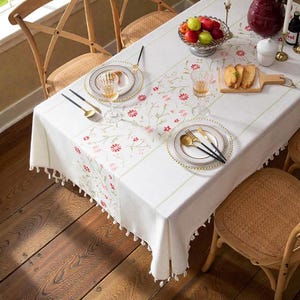 Può includere: Una tovaglia bianca con un motivo floreale, apparecchiata per un pasto. La tavola è apparecchiata con piatti, posate e bicchieri. Una ciotola di frutta e un tagliere di legno sono anche sulla tavola. La tovaglia ha un bordo con frange.