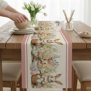 May include: A beige Easter table runner with a repeating pattern of bunnies, baskets, and eggs. The runner has a pink border with vertical stripes. The table is set with white plates, a wooden bowl, and a vase of white tulips.