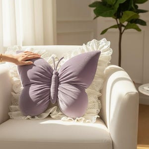 Puede incluir: Un cojín con forma de mariposa lavanda con un borde de encaje blanco descansa sobre un sillón blanco. Las alas del cojín tienen vetas sutiles, y el cuerpo es de un tono lavanda más oscuro. La estética general es suave y decorativa.