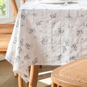 Könnte beinhalten: Weißes Tischtuch mit floralem Muster. Der Stoff weist ein sich wiederholendes Design aus kleinen, dunkelgrünen und weißen Blumenclustern auf. Das Tischtuch ist über einen Holztisch drapiert, mit einem Blick auf einen Stuhl im Vordergrund.