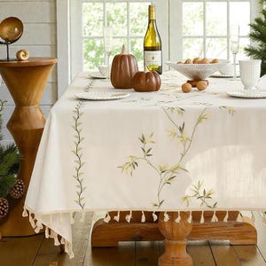 May include: A cream-colored tablecloth with embroidered green and yellow floral designs and a tassel trim. The table is set with white plates, glasses, and brown pumpkin decorations. A bottle of wine is also on the table.