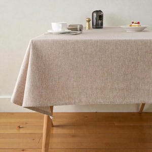 Op de afbeelding: Een beige linnen tafelkleed bedekt een houten tafel, met een witte kop en schotel, een klein bordje met eten en een zout- en peperstel. Het tafelkleed heeft een textuur en hangt over de randen van de tafel.