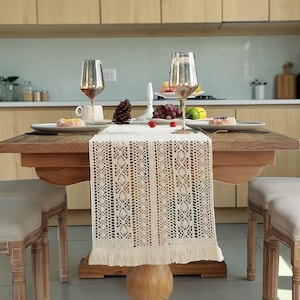 May include: A dining table set for two with a cream-colored, open-weave table runner. Two wine glasses, plates with food, and a pinecone decorate the table. The table is made of wood and has a pedestal base.