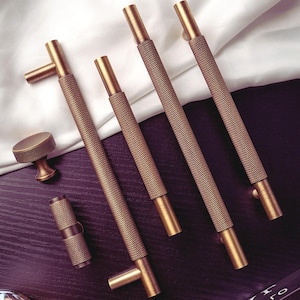 Pode incluir: Uma coleção de ferragens de armário em latão antigo, incluindo várias puxadores de barra texturizados, um botão redondo e um pequeno puxador cilíndrico. As ferragens estão dispostas sobre uma superfície escura com um pano branco ao fundo.