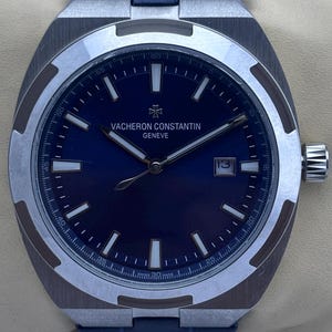 Könnte beinhalten: Eine luxuriöse Armbanduhr mit dunkelblauem Zifferblatt und silberfarbener Lünette. Die Uhr trägt den Markennamen "VACHERON CONSTANTIN GENEVE" und eine Datumsanzeige. Das Armband ist aus blauem Leder.