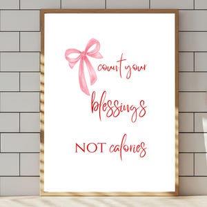 May include: A framed print with a white background features a pink watercolor bow and red script text. The text reads "count your blessings not calories". The frame is a light brown color.