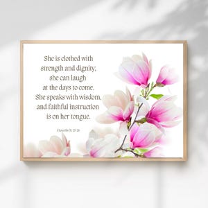 Puede incluir: Una impresión enmarcada con un arreglo floral de flores de magnolia rosas y blancas. La obra de arte incluye el texto "Se reviste de fuerza y dignidad, y se ríe sin temor al futuro. Abre su boca con sabiduría, y la instrucción de la bondad está en su lengua." Proverbios 31:25-26.