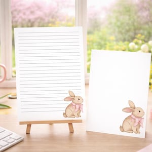 May include: Two white notepads with a watercolor illustration of a bunny wearing a pink bow. One notepad is on a small wooden easel. A pink mug with pencils, a pink tulip, and a keyboard are on the desk.