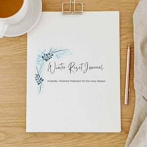 Puede incluir: Un diario blanco con el texto "Winter Reset Journal" y el subtítulo "A Gentle, Feminine Reflection for the Cozy Season". El diario está sujeto por un clip plateado. Una taza de té y un bolígrafo también están sobre la superficie de madera.