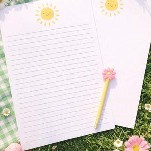 May include: Two white lined stationery sheets with a smiling sun graphic at the top. A yellow pen with a pink flower topper rests on the paper. The scene is set on a green checkered cloth and grass, with daisies and Easter eggs.