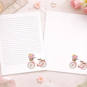 May include: Two sheets of white stationery, one lined and one blank, feature a pink bicycle with a basket of flowers. The stationery is decorated with pink flowers and pencils, creating a floral theme.