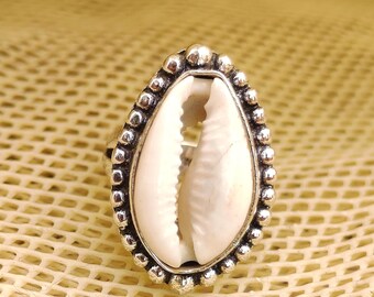Könnte beinhalten: Ein silberner Ring mit einem weißen Muschelschalen-Mittelstück. Die Schale ist oval und in einen perlenbesetzten, tropfenförmigen Rahmen gefasst. Der Ring wird auf einer strukturierten, hellgelben Oberfläche gezeigt.
