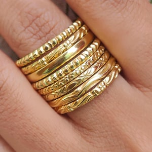 Puede incluir: Primer plano de un dedo adornado con múltiples anillos dorados. Los anillos muestran diversas texturas, como superficies lisas, punteadas y estampadas. Los anillos están hechos de un material metálico brillante.