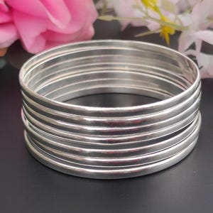 Könnte beinhalten: Ein Stapel von sieben silbernen Armreifen. Die Armreifen sind glatt und glänzend und reflektieren das Licht. Sie sind übereinander gestapelt, wodurch ein geschichteter Effekt entsteht. Im Hintergrund sind rosa und weiße Blumen zu sehen.