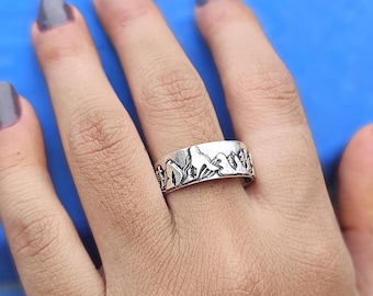 Puede incluir: Un anillo plateado con un diseño de cordillera. El anillo presenta un paisaje detallado de montañas y árboles. El anillo se lleva en un dedo sobre un fondo azul.