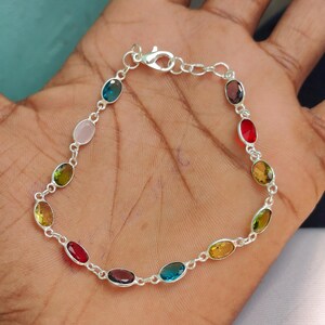 Puede incluir: Una pulsera de plata con una serie de piedras preciosas ovaladas en varios colores, incluyendo azul, rosa, verde, amarillo y rojo. La pulsera se muestra sobre un fondo oscuro, resaltando los colores vibrantes de las piedras.