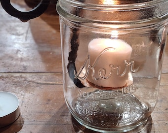 May include: A clear glass Kerr self-sealing mason jar containing a small white tea light candle. A black, hand-forged metal S-hook is hooked over the rim of the jar, extending downward into the interior to hold the candle in place.