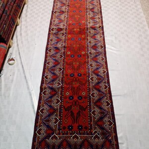 May include: A long, rectangular red rug with intricate geometric and floral patterns in blue, brown, and white. The rug features a central design with repeating motifs and a detailed border. The rug is displayed on a white surface.