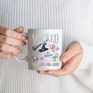 May include: White ceramic mug with the word "WICKED" in iridescent lettering, along with illustrations of a witch's hat, broom, and other magical elements. The phrase "CHANGED FOR GOOD" is printed below the illustrations.