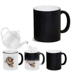 May include: A collection of black and white heat-sensitive mugs. One mug features a cat peeking through a torn paper effect. The black mugs turn white to reveal the cat image when hot liquid is added. A white teapot is also present.