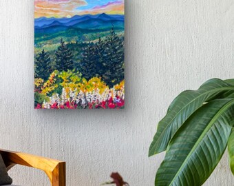 May include: A landscape painting showing a mountain scene at sunset. The artwork features layers of blue mountains, green trees, and colourful wildflowers in the foreground. The sky transitions from orange to blue.