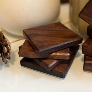 May include: A stack of square wooden coasters. The coasters are made of dark brown wood with a visible grain. The edges are beveled, and the coasters are stacked on top of each other.